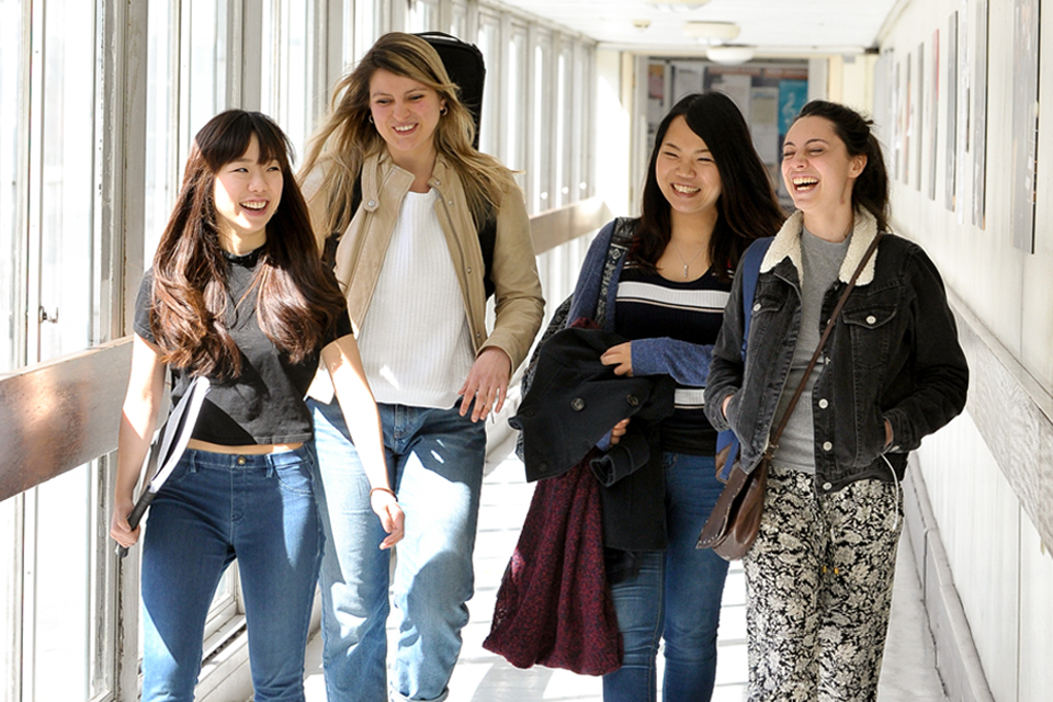 A group of female students, holding music sheets, smiling and chatting while walking in a well-lit corridor at the RCM.